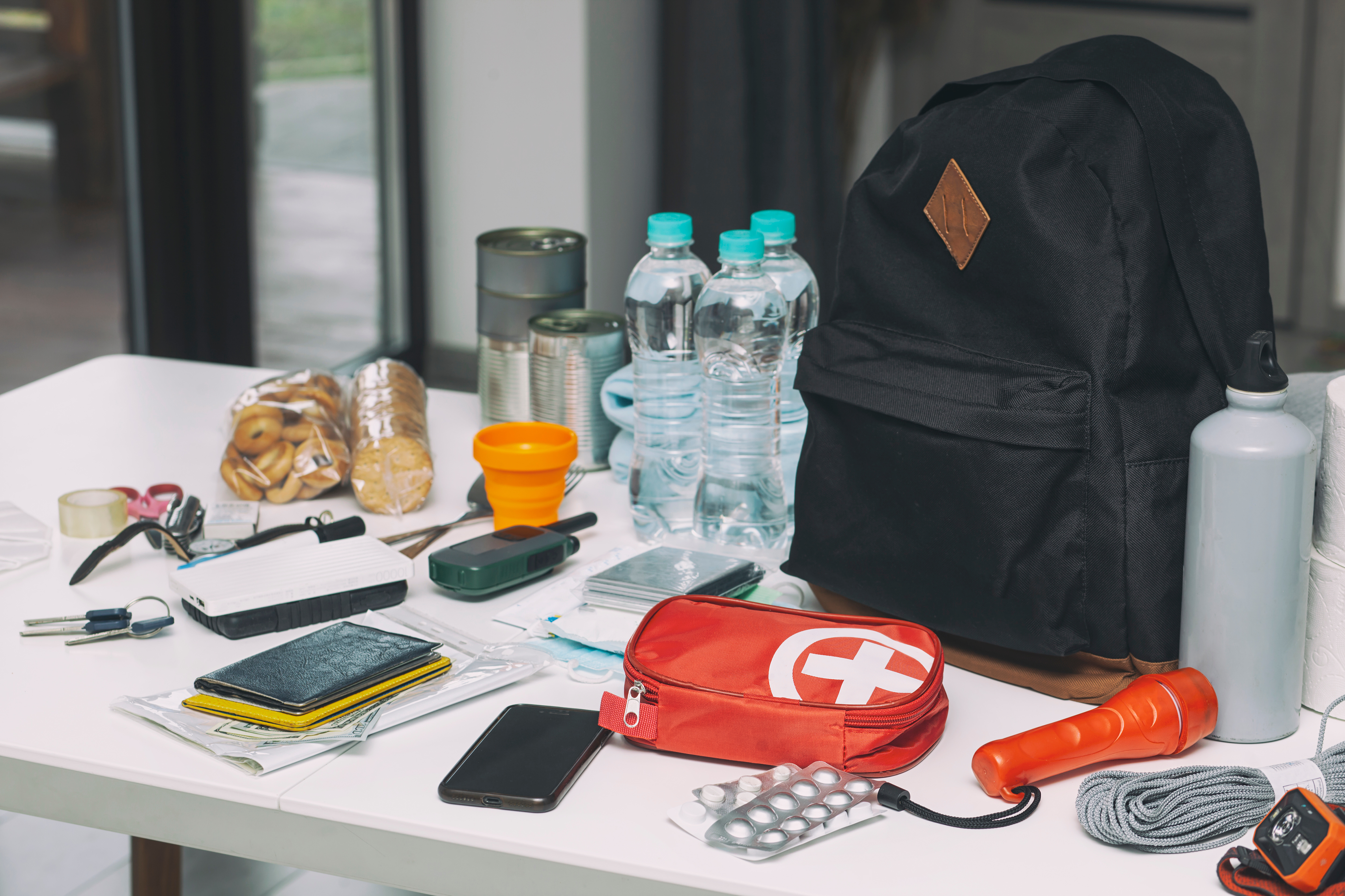 Emergency supplies arranged on a table, including bottled water, canned food, a backpack, first aid kit, flashlight, phone, medication and other essential items.