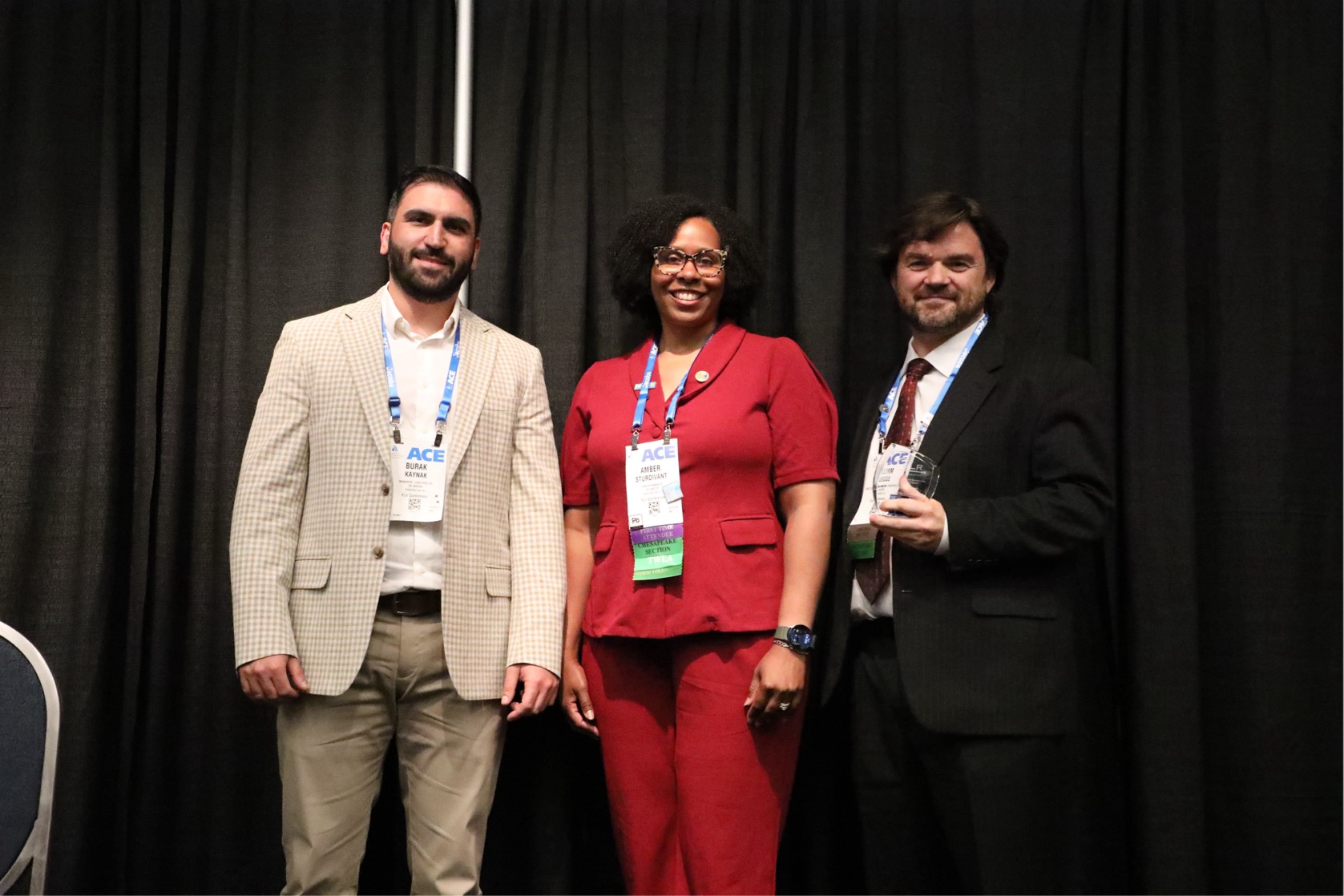 Burak Kaynak, Amber A. Sturdivant, and William Elledge pose together onstage at the American Water Works Association’s ACE26 conference. Amber A. Sturdivant stands between Burak Kaynak and William Elledge, who holds an award recognizing Lead Free DC’s outstanding leadership in lead service line replacement. All three wear conference badges and smile in front of a black curtain backdrop.