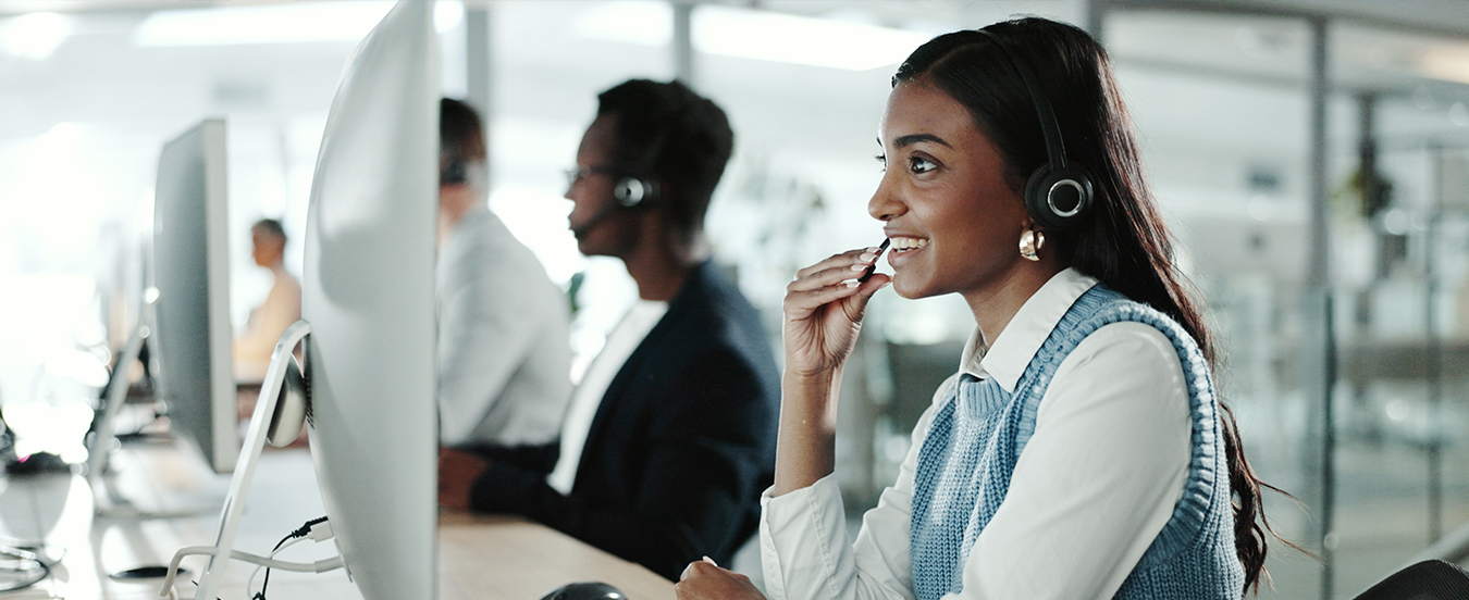 A customer care associate speaks by phone to a customer. The associate is holding a microphone in her right hand while she looks at a computer screen.