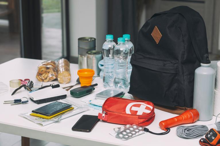 Emergency supplies arranged on a table, including bottled water, canned food, a backpack, first aid kit, flashlight, phone, medication and other essential items.