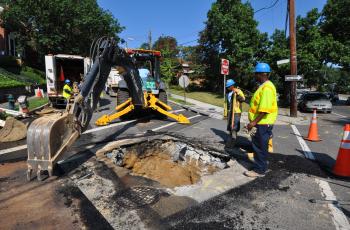 Two workers making a water main break repair with a excavator nearby